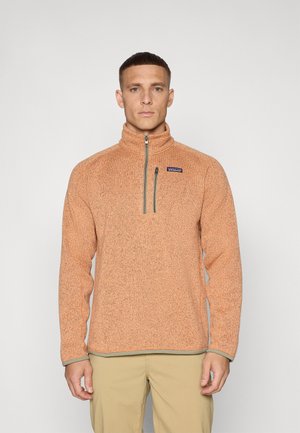 Fleecepullover i ljusorange med zip-krage, kontrasterande grön dragkedja och en logotypapplikation på bröstet. Texturerad yta.