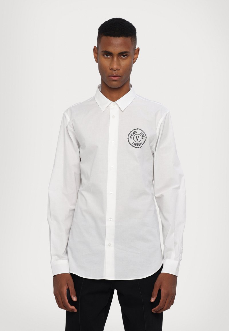 Biała koszula z długim rękawem i kołnierzykiem zapinanym na guziki, z okrągłą czarną łatą z tekstem i logo z lewej strony. Gładka tekstura materiału.