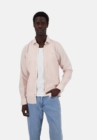 Camicia a maniche lunghe a righe in pesca chiaro e crema, con bottoni e una morbida texture in cotone, indossata sopra un canottiera bianca aderente, abbinata a jeans azzurri chiari.