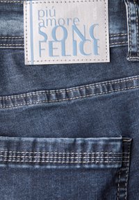 Tissu en denim bleu foncé avec une surface texturée, doté d'une étiquette en cuir cousue, sur laquelle est inscrit "più amore SONO FELICE" en bleu clair.