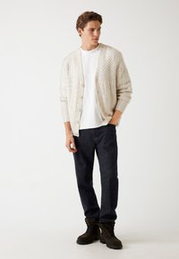Cremfarbener, gestrickter Cardigan mit einem strukturierten Muster, Knopfverschluss, kombiniert mit einem weißen T-Shirt und dunklen Jeans. Braune Stiefel vervollständigen das Outfit.