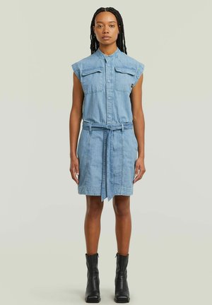 Femme aux cheveux tressés portant une robe en jean bleu clair sans manches avec poches avant et ceinture, associée à des bottines noires, debout face à l'avant.
