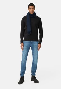 Zwarte gebreide trui, donkerblauwe sjaal met franjes, lichtblauwe jeans, zwarte enkellaarzen. Model staat tegen een effen witte achtergrond.