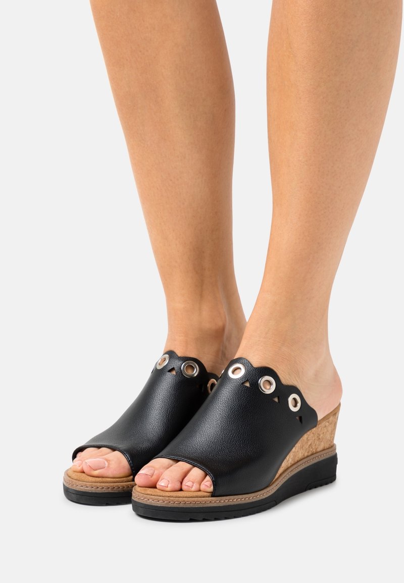 Pieds portant des sandales compensées noires à bout ouvert avec des bords ondulés et des œillets en métal, posés sur un fond gris clair.