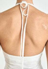 Vestido blanco sin mangas con cuello atado, que presenta una textura suave y un cuerpo ajustado. Muestra una espalda elegante con sutiles detalles de piel.