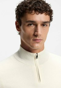 Cremefarbener Strickpullover mit hohem Kragen und Viertel-Reißverschluss. Verfügt über eine gerippte Textur und minimalistisches Design.