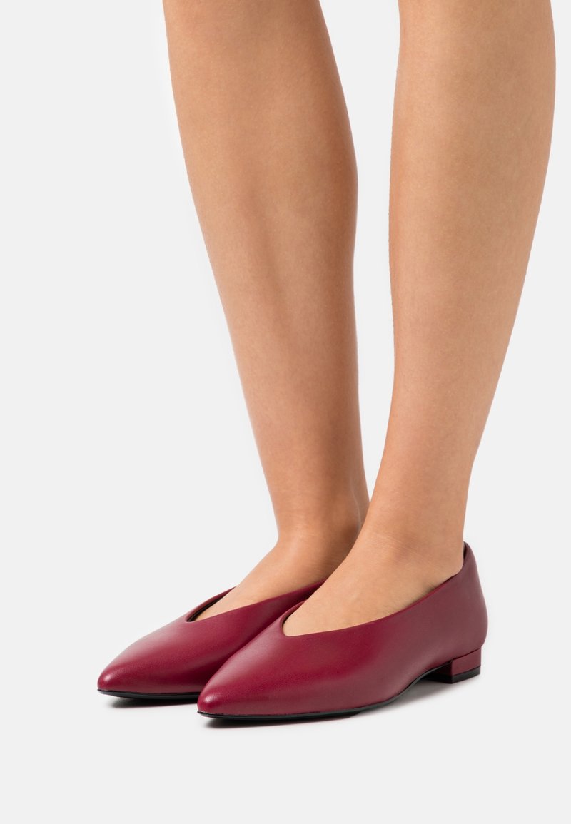 Ballerines en cuir bordeaux avec un bout pointu, un petit talon et un design découpé sur les côtés, présentant une texture lisse et un matériel minimal.
