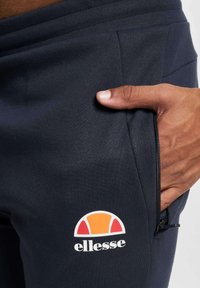 Joggers azul marino hechos de una tela suave presentan un corte ajustado, un bolsillo con cremallera y una etiqueta de logo en naranja y rojo cerca de la cadera.