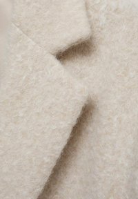 Beige villakangastakki, jossa on rakenne ja pehmeä pinta, jossa on lovettu kaulus ja siistit linjat, jotka korostavat pörröistä ja kevyttä ilmettä.