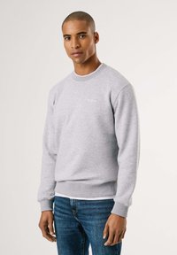 Sudadera gris con puños y dobladillo acanalados, con cuello redondo y pequeño logo blanco en el pecho. Combinada con jeans azules.
