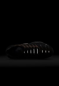 Chaussure de sport noire avec des accents bruns, présentant un dessus texturé, des détails réfléchissants sur le système de laçage et une conception à bout rond.