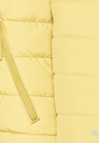 Sección de chaleco acolchado amarillo con textura acolchada, costuras verticales y cierre de cremallera. Tela suave con un acabado mate.