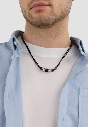 Primer plano de un hombre que lleva un collar de cuerda negra con cuentas cilíndricas plateadas sobre una camiseta blanca y una camisa azul claro abotonada y abierta.