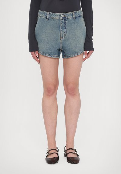 Shorts en denim bleu clair avec taille haute. Présentent un bouton et une fermeture éclair à l'avant, des ourlets roulés et une coupe standard, associés à des chaussures sombres.