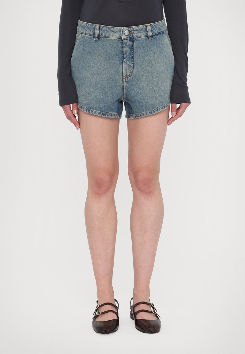 Pantalones cortos de mezclilla en azul claro con una cintura alta. Presentan un botón y cremallera en la parte delantera, dobladillos enrollados y un ajuste estándar, combinados con zapatos oscuros.