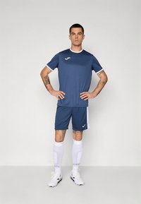 Tamsiai mėlynos spalvos sportinis marškinėlių ir šortų komplektas, pagamintas iš lengvos medžiagos. Yra balta apdaila, logotipas ant krūtinės ir baltos kojinės iki kelių.