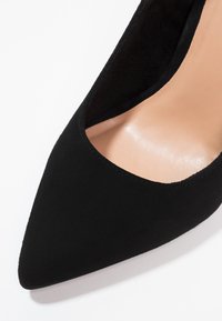 Escarpin noir en daim à bout pointu avec doublure beige, vu de dessus sur un fond blanc.