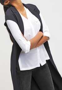 Lång marinblå väst över en vit skjorta med knappar och uppvikta ärmar, tillsammans med mörka jeans. Skjorta har två bröstfickor.