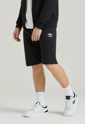 Pantaloni corti sportivi neri con vita elastica, caratterizzati dal logo Umbro bianco. Abbinati a sneaker bianche e calze bianche a coste. Texture liscia.