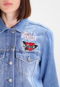 Denimjacka i ljusblått med slitna detaljer. Har en röd hjärtapplikation med texten "WILD HEART" och gröna blad på v änster bröst.