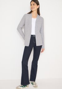 Šedý žebrovaný kardigan přes bílý top, doplněný tmavými rozšířenými džínami a teniskami se zelenými akcenty. Jednoduchý design s otevřeným předním dílem.