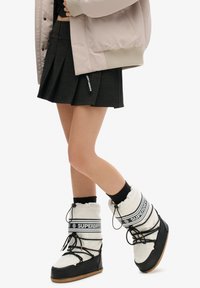 Bottes de neige blanches et noires avec des revers rembourrés et des lacets, associées à une jupe plissée noire et une veste bomber rose pâle avec des poignets côtelés.