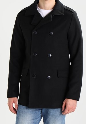 Homme portant un caban noir à double boutonnage avec six boutons et des poches à rabat, associé à un jean bleu clair et une chemise blanche.