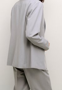 Costume gris sur mesure comprenant un blazer à simple boutonnage avec une coupe droite et un pantalon assorti, fabriqué à partir d'un tissu lisse et léger.