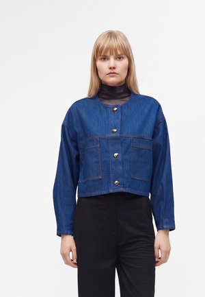 Femme aux cheveux blonds et raides porte une veste courte en jean bleu avec des boutons dorés par-dessus un top noir transparent et un pantalon noir.
