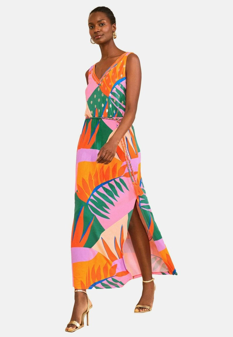 Frau trägt ärmelloses, buntes Maxikleid mit abstrakten orange-, grünen, pinken und violetten Blattmustern und Seitenschlitz, kombiniert mit goldenen Absätzen.
