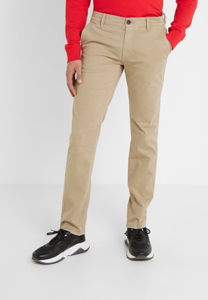 Man draagt beige chino's, zwarte sneakers en een rood shirt met lange mouwen, staat met één hand in zijn zak tegen een witte achtergrond.