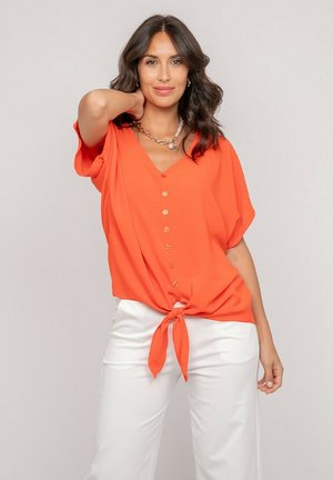 Femme aux cheveux ondulés foncés portant un chemisier orange boutonné noué à la taille et un pantalon blanc, posant avec une main derrière le cou.