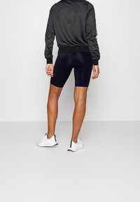 Personne debout de dos portant un short de cyclisme noir, une veste noire avec des manches à motifs, et des baskets blanches avec des accents néon.