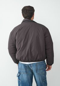 Gepolsterte, dunkelgraue Jacke mit hohem Kragen und elastischen Bündchen. Getragen über einem weißen Hemd, kombiniert mit hellblauen Cargo-Jeans.