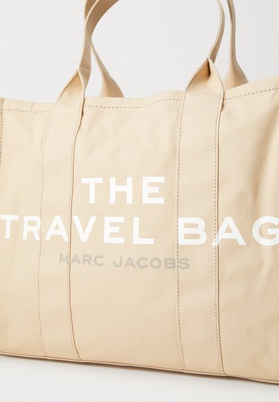 Bež platnena torba s dvije čvrste ručke. Ima dominantne bijele natpise: "TORBA ZA PUTOVANJE" i "MARC JACOBS" u sivoj boji. Šavovi su vidljivi.