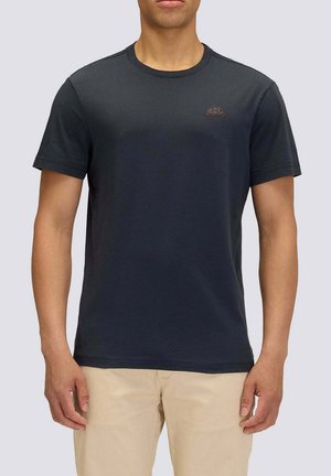 Uomo che indossa una t-shirt semplice blu navy scuro a maniche corte con un piccolo logo sul petto e pantaloni beige chiaro su sfondo grigio.
