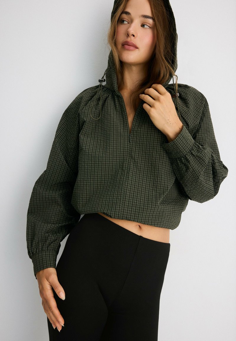 Jeune femme portant un sweat à capuche court à carreaux vert foncé et des leggings noirs taille haute, regardant sur le côté avec une main près du visage.