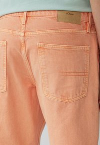 Pantalons en denim couleur pêche avec une texture lisse, présentant deux poches arrière et une étiquette en cuir portant le nom "s.Oliver". Détails de coutures simples.