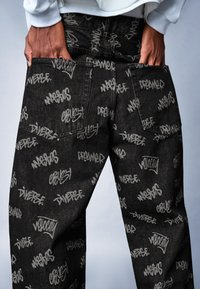 Černé džínové kalhoty s různorodým šedým textovým potiskem, velkými kapsami a volným střihem. Látka má jemnou texturu.