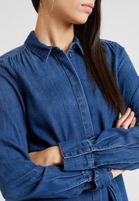 Camicia in denim blu scuro, con colletto e chiusura a bottoni. Maniche lunghe con polsini chiusi da bottoni e leggero arricciamento alle spalle. Texture liscia.