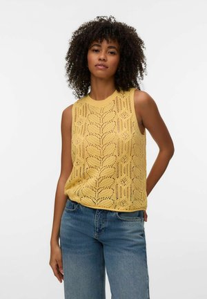 Mujer con cabello rizado que lleva una camiseta sin mangas de punto amarilla y jeans azules, de pie frente a un fondo claro y liso.