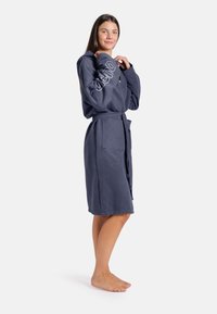 Námořnicky modrý koupací plášť s kapucí vyrobený z měkké tkaniny, s páskem v pase, kapsami a bílým textovým detailem na rukávu.