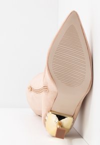 Beige mocka högklackad sko med spetsig tå, guldaccenter vid klacken och texturerad sula för grepp. Dragkedjedetalj på sidan.