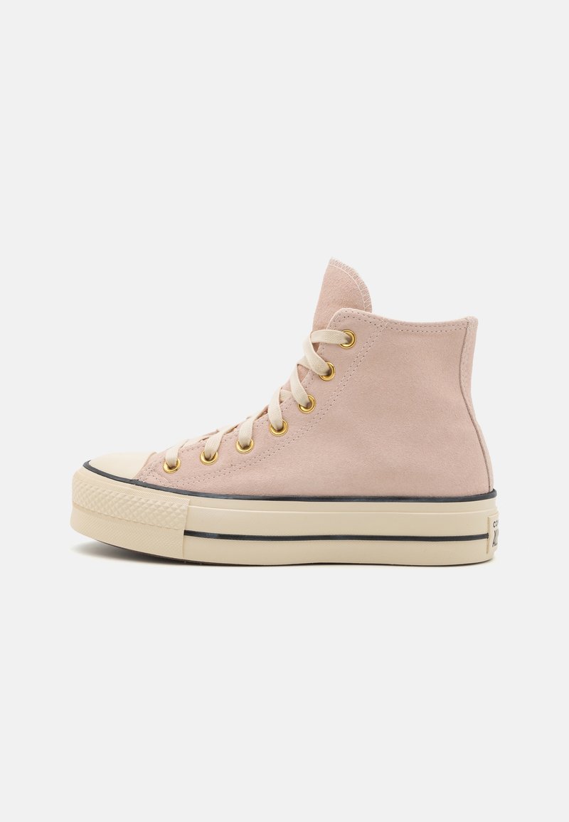 Wysokie beżowe zamszowe sneakersy z kremową gumową podeszwą, czarnym paskiem, złotymi oczkami i kremowymi sznurowadłami na białym tle.