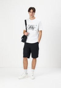 Biely tričko s grafickým dizajnom, čierne cargo šortky, biele tenisky a malá čierna crossbody taška, umiestnené na neutrálnom pozadí.