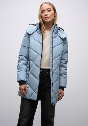 Hellblauer Puffer-Mantel mit diamantförmiger Steppung, verstellbare Kapuze, Reißverschluss und schwarzen Ärmelakzenten, getragen über einem cremfarbenen Pullover und dunklen Jeans.