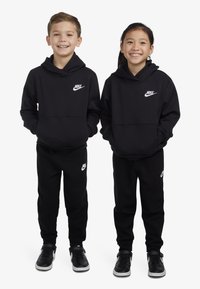 Fekete Nike kapucnis pulóver és melegítőnadrág fehér logóval. Mindkét öltözék laza szabású, puha anyagú és zsinórral ellátott kapucnis. Cipőkkel hangsúlyozva.