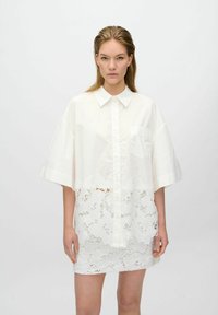 Robe chemise blanche oversize avec un col pointu, présentant un panneau inférieur en dentelle. Matière en coton doux avec des manches courtes et une poche poitrine.