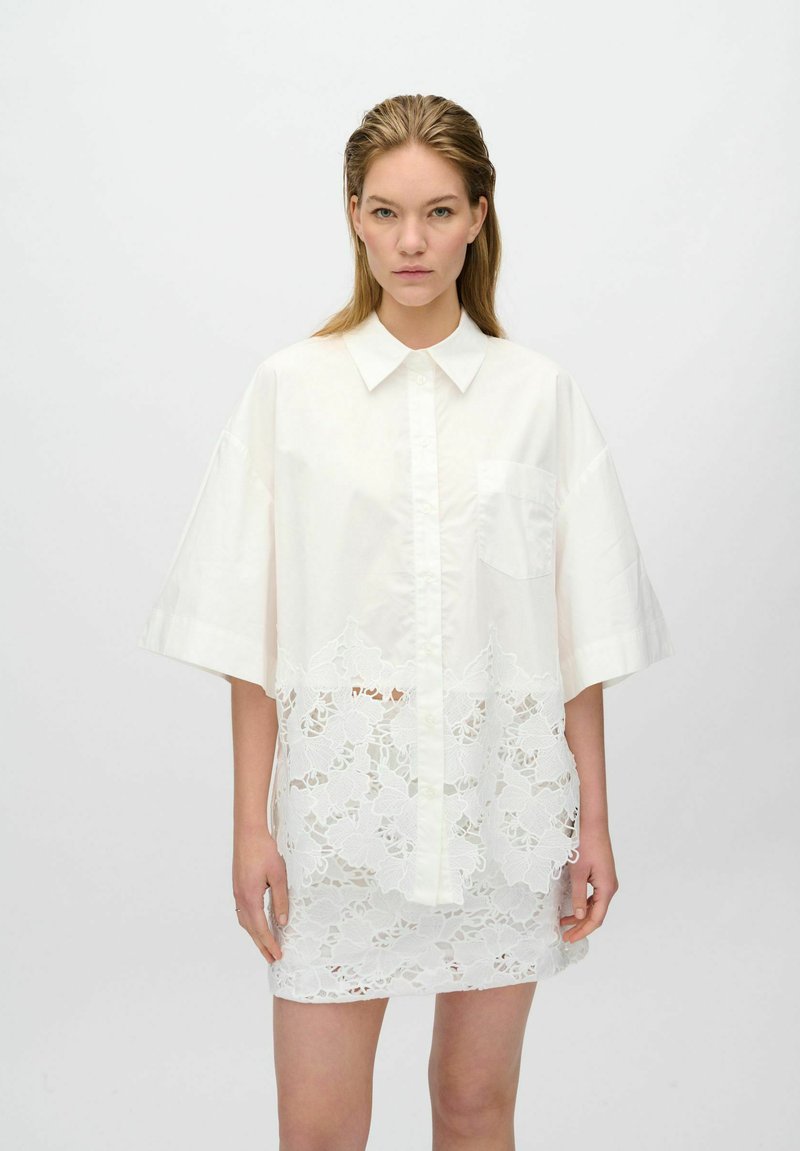Robe chemise blanche oversize avec un col pointu, présentant un panneau inférieur en dentelle. Matière en coton doux avec des manches courtes et une poche poitrine.