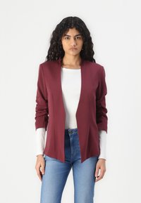 Burgundský blazer s otevřeným předním designem, dlouhými rukávy a vystuženými rameny, vrstvený přes bílou košili s dlouhým rukávem. Texturovaná tkanina.